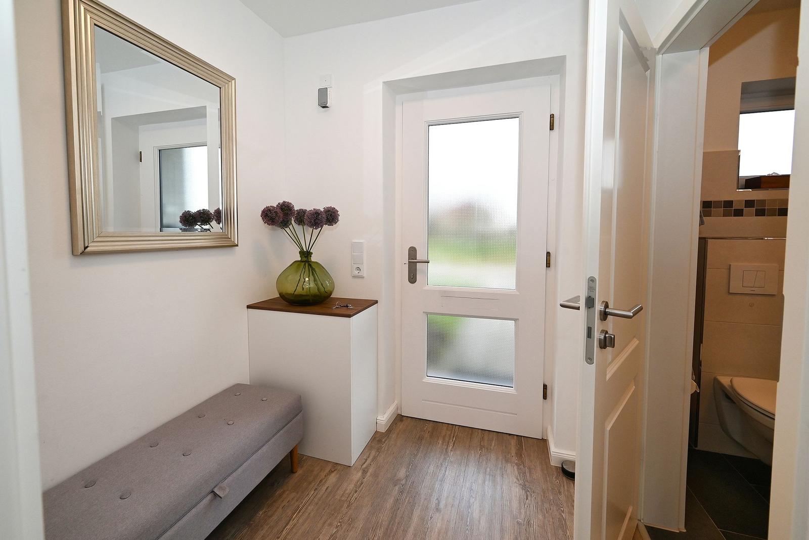 Eingangsbereich mit Spiegel, Sitzbank und Tür. Nebenraum ist Bad mit Toilette.