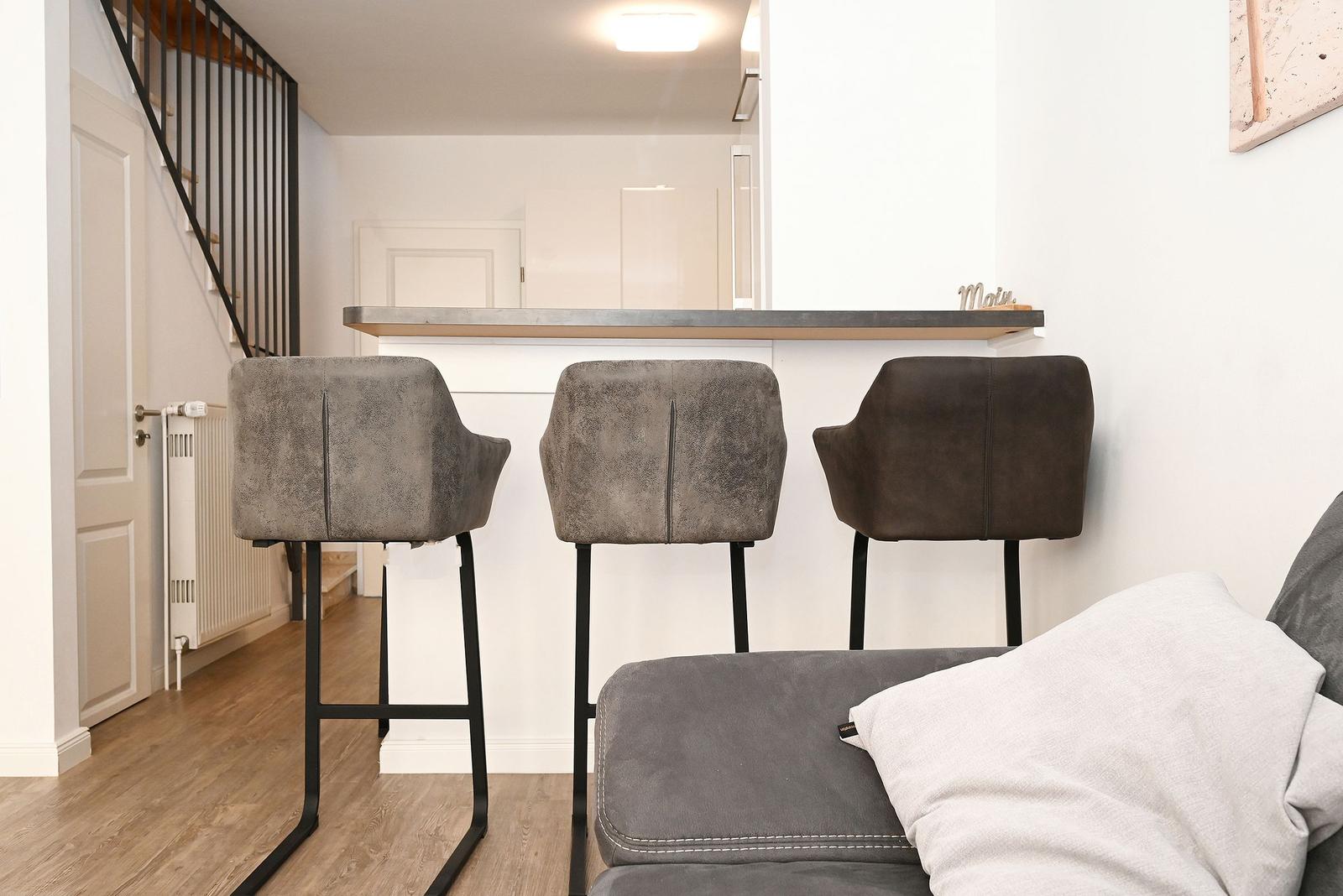 Barhocker vor einer Küchenzeile mit Tresen. Im Hintergrund Treppe und Wohnbereich.