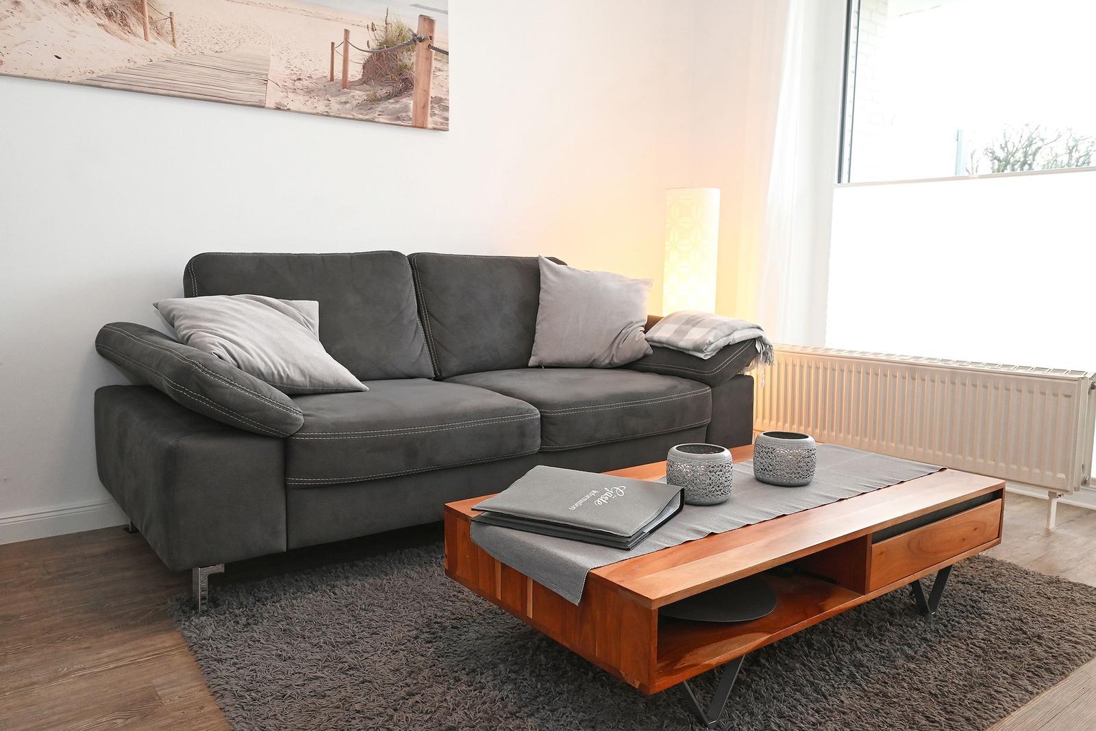 Ein grauer Sofa mit Kissen steht vor einem Fenster. Davor befindet sich ein Holztisch.