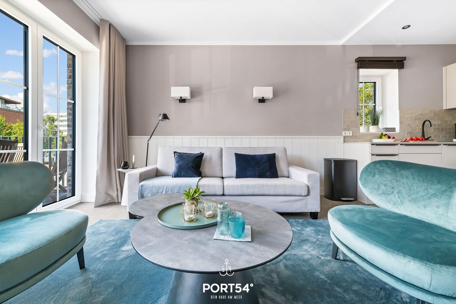 Wohnbereich mit Sofa, zwei Sesseln und runden Tisch. Küche und Balkon sind sichtbar.