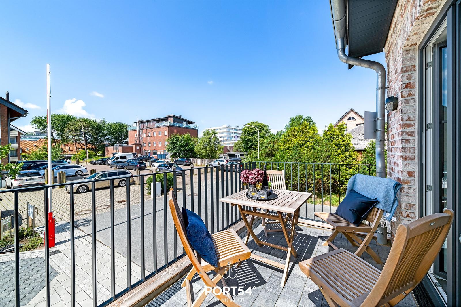 Terrasse mit Holztisch und Stühlen, Blick auf Parkplatz und Gebäude.