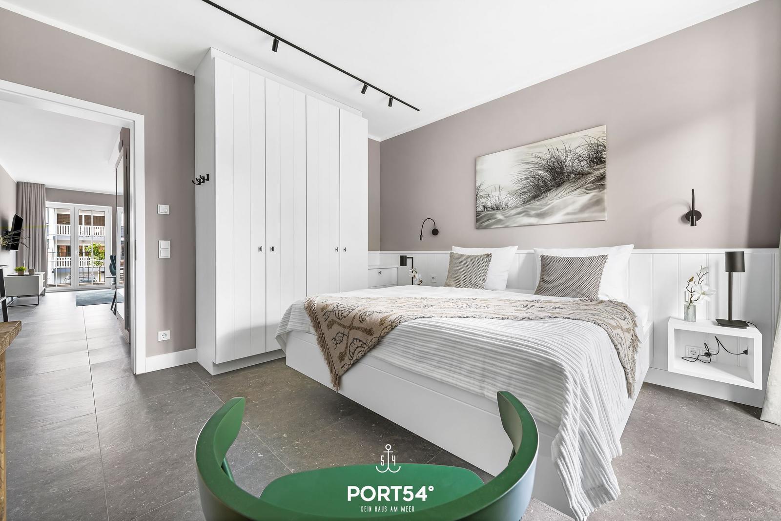 Schlafzimmer mit weißem Bett, Kleiderschrank und Wandbild. Durchgang zu Wohnbereich mit Balkon.