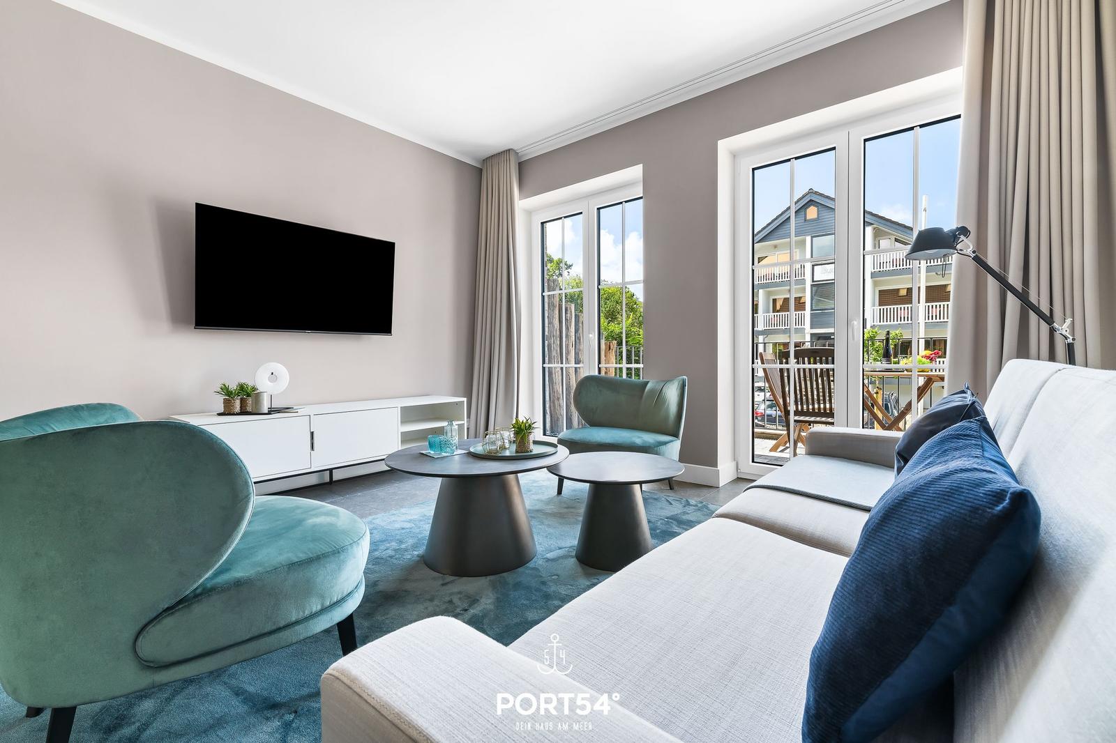 Wohnzimmer mit Sofa, Sesseln, Couchtisch und TV. Durchgang zur Terrasse mit Blick auf Gebäude.