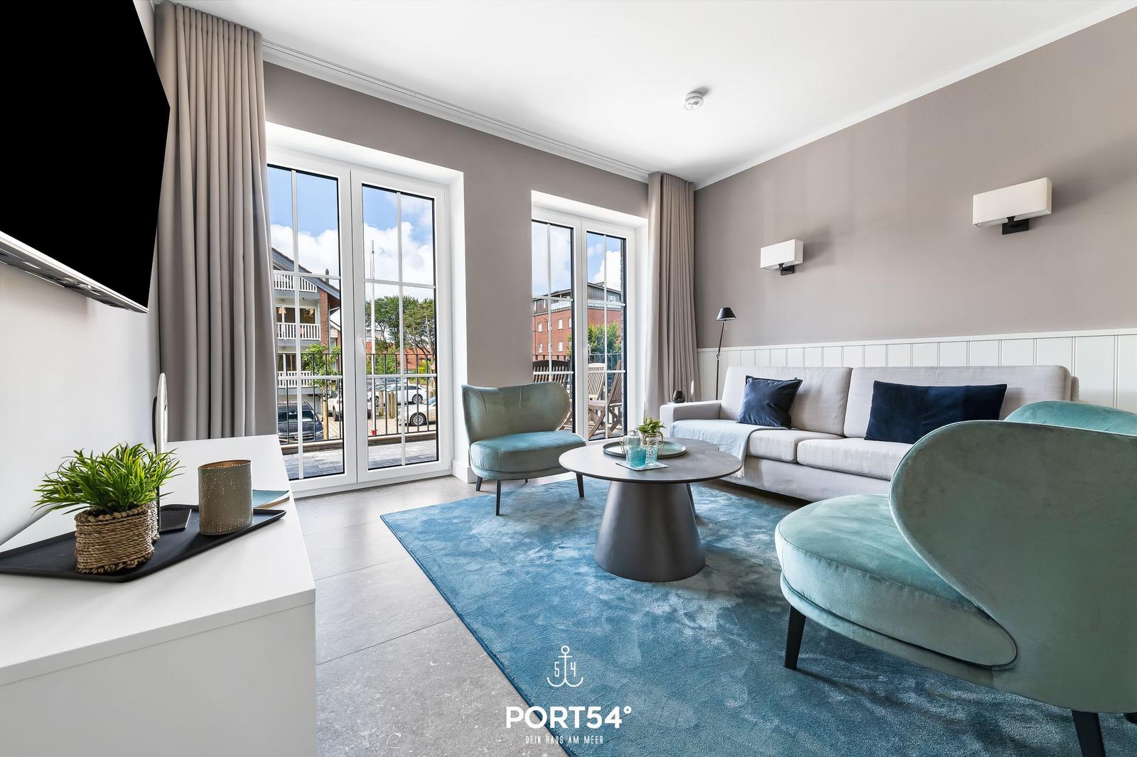 Wohnzimmer mit Sofa, Sesseln und TV. Durchgänge zu Balkon. Blauer Teppich.
