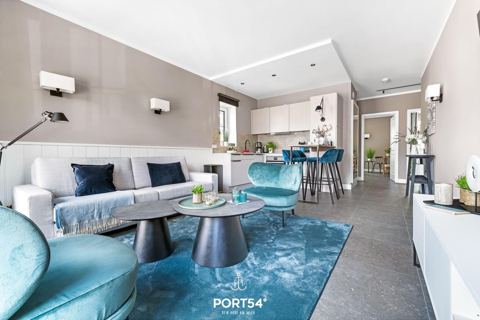 Wohnbereich mit Sofa, Sessel und Küchenbereich. Blauer Teppich und moderne Einrichtung.
