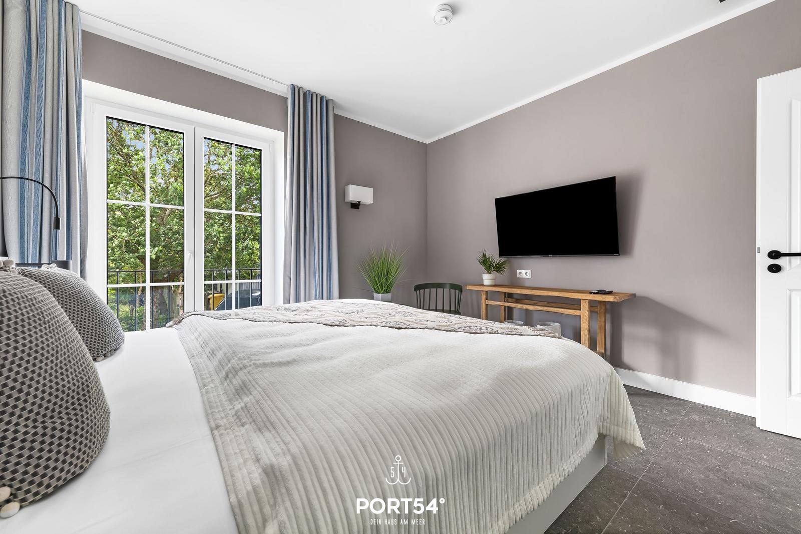 Schlafzimmer mit Bett, TV und Balkon mit Blick auf Bäume.