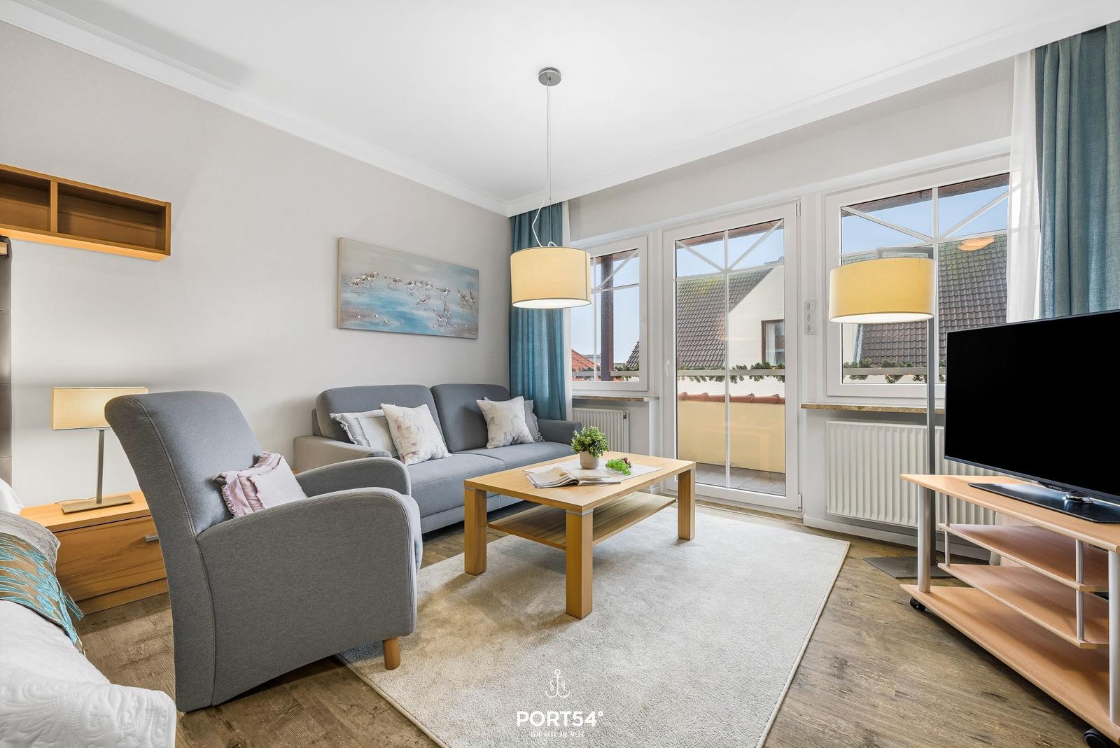 Wohnzimmer mit Sofa, Sessel, Couchtisch und TV. Terrasse über Schiebetür.