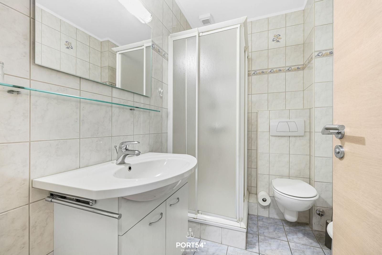 Badezimmer mit Dusche, Waschbecken und Toilette. Fliesenwand mit Muschel-Motiv.