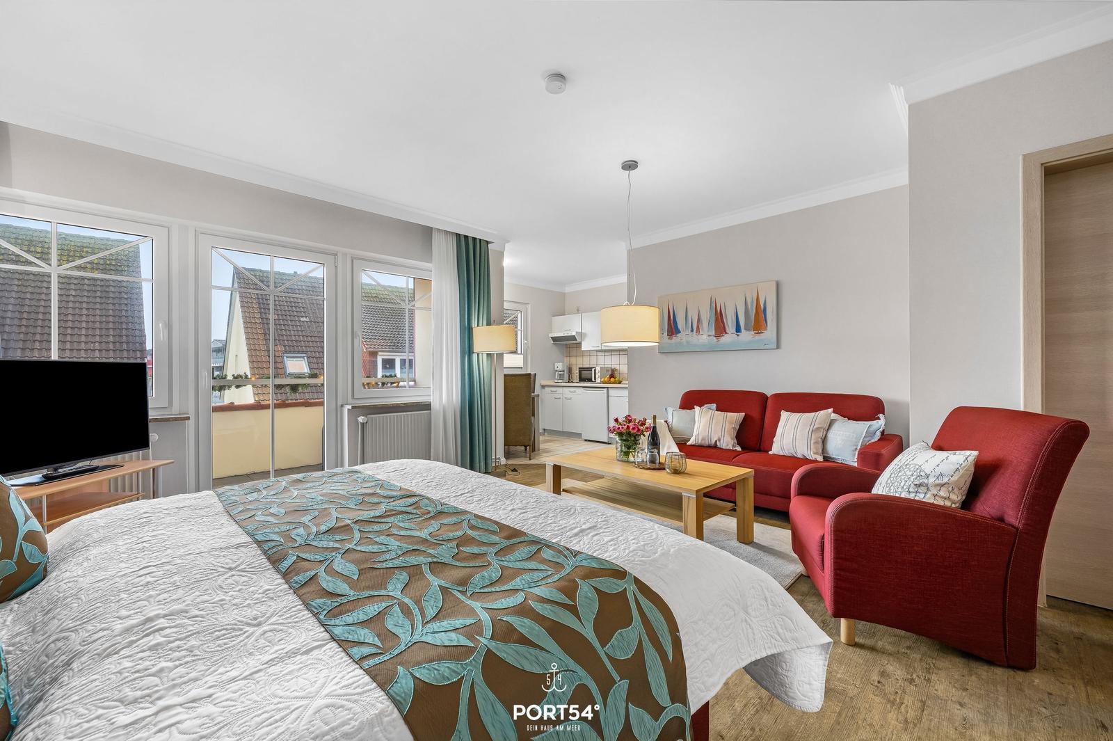 Ein Schlafzimmer mit Blick auf Balkon, Wohnbereich mit rotem Sofa und Küchenbereich.