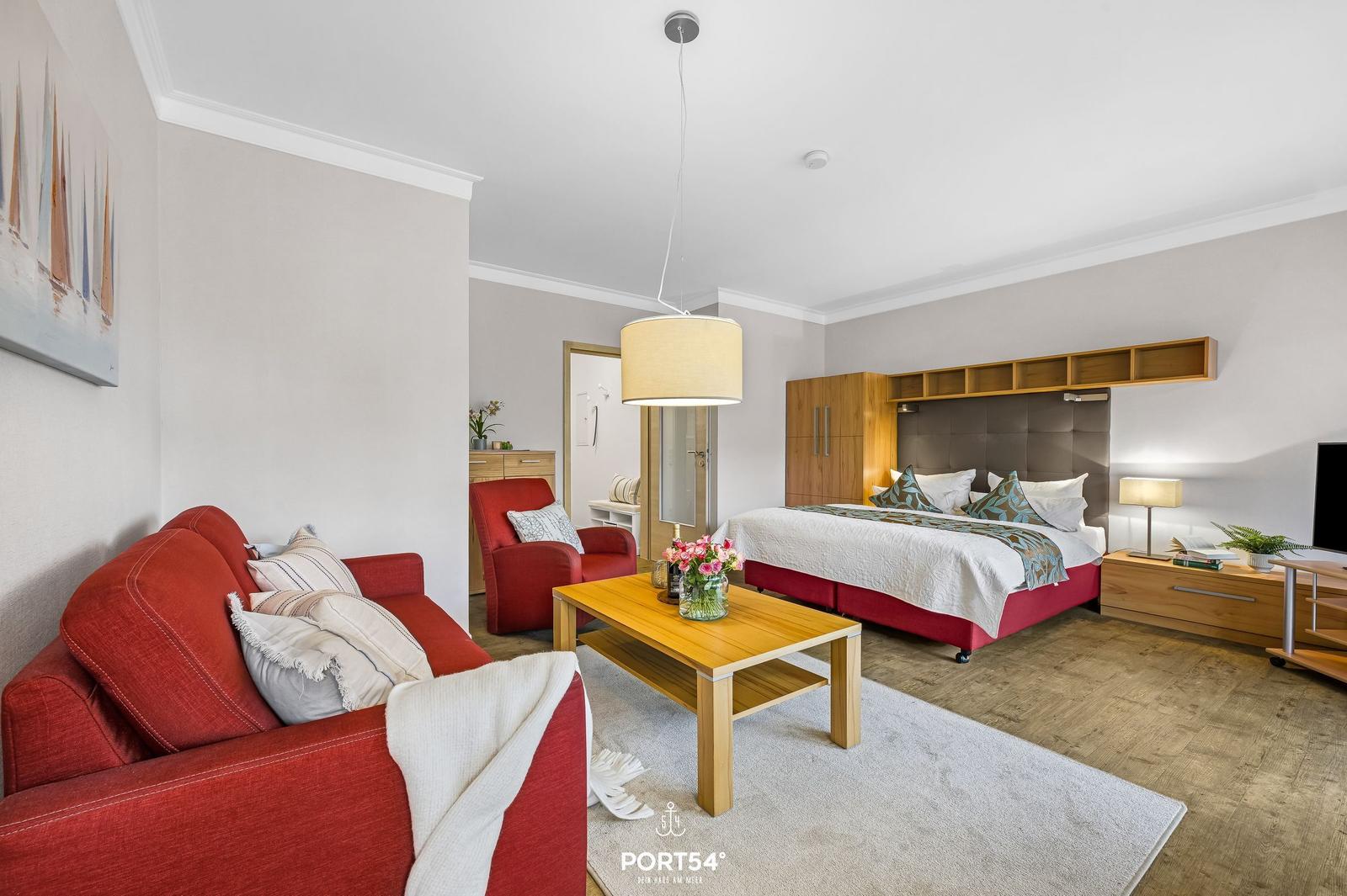 Wohn- und Schlafbereich mit rotem Sofa, Holztisch und Bett mit Holzgestell.