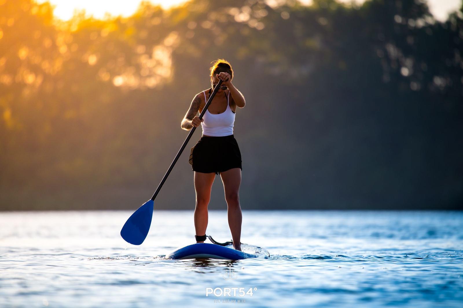 Eine Person steht auf einem Stand-Up-Paddleboard auf dem Wasser und rudert mit einem blauen Ruder.