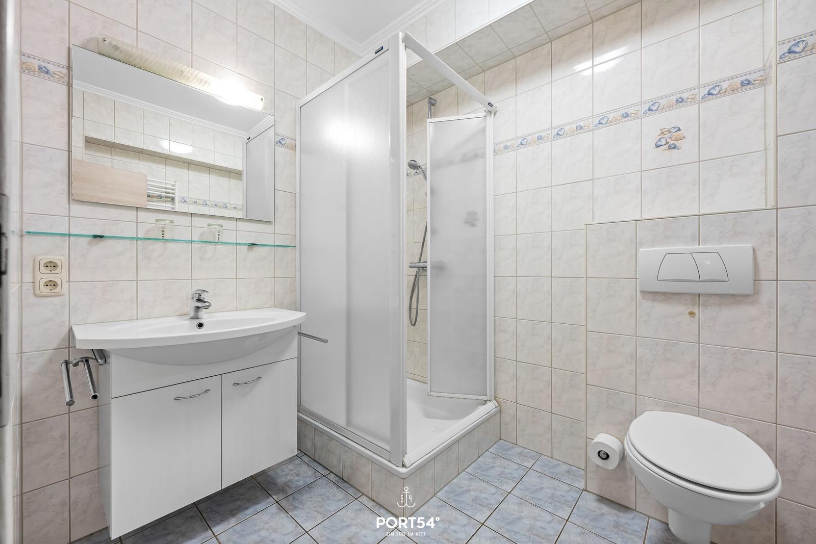 Badezimmer mit Dusche, Waschbecken und Toilette. Fliesenwand mit Muschelrand.