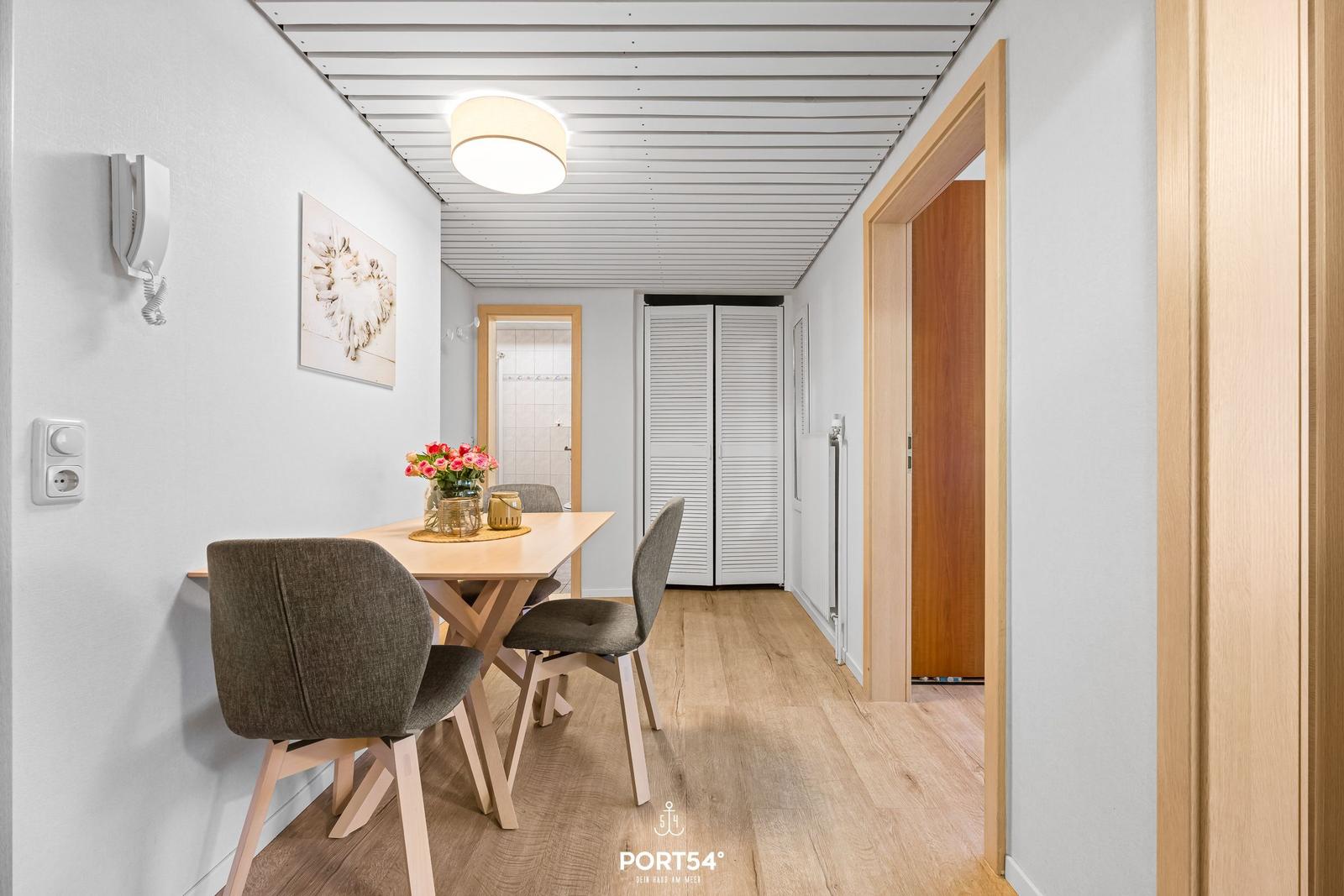 Essbereich mit Tisch, Stühlen und Blumen. Hintergrund: Bad und Schrank.