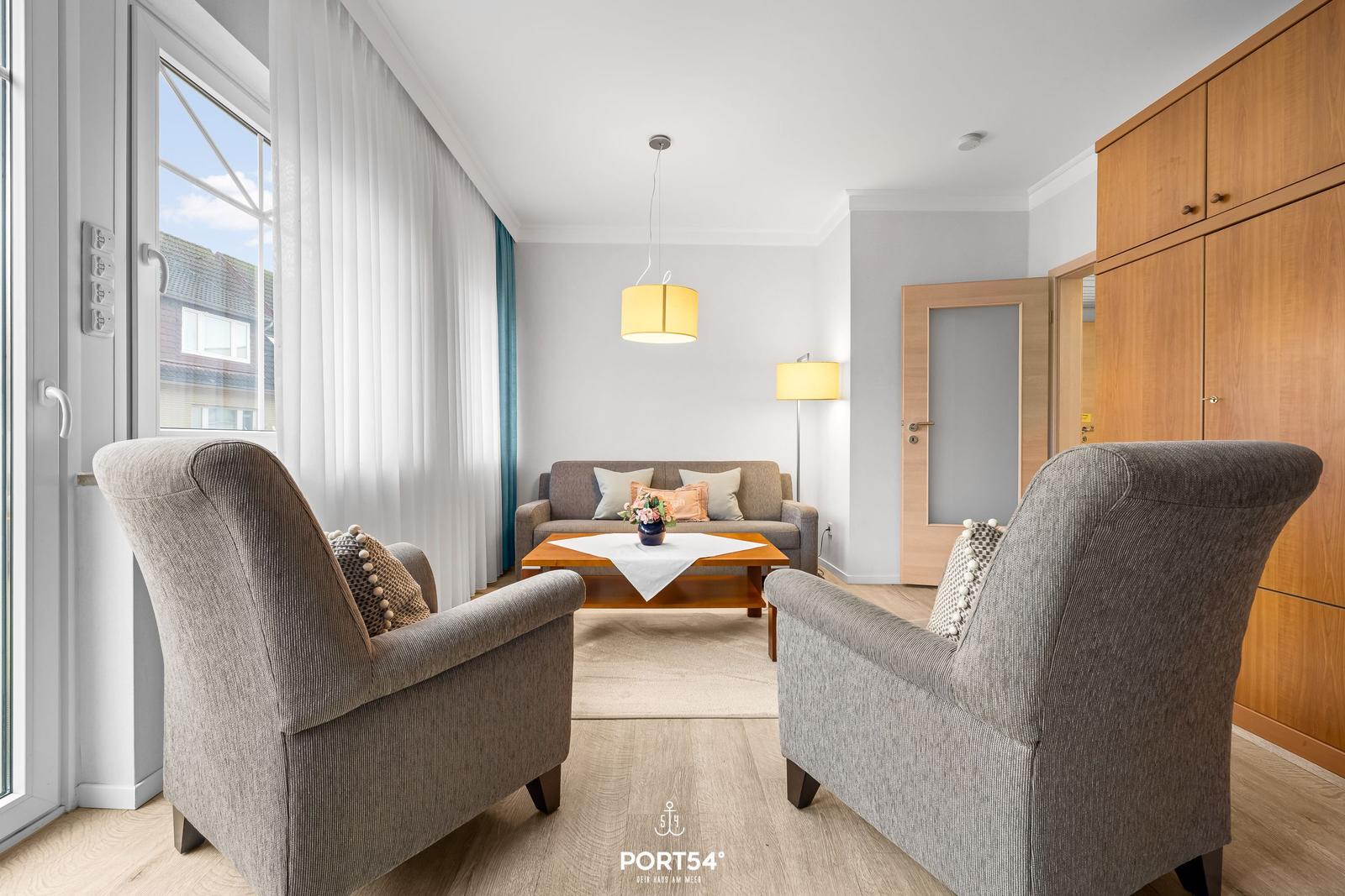 Wohnzimmer mit zwei Sesseln, Sofa, Tisch und Fenster mit Blick nach außen.