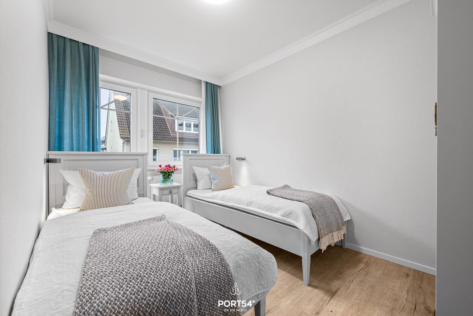 Zwei Einzelbetten mit weißen Bettwäschen und grauen Decken. Fenster mit blauen Vorhängen.