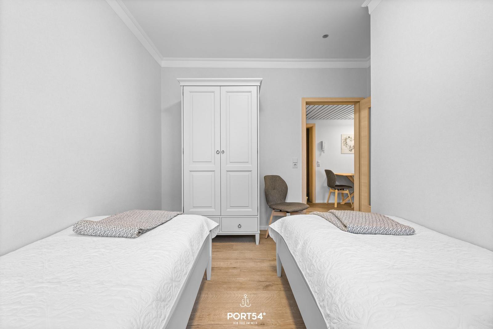 Zwei Betten, Kleiderschrank und Stuhl im Schlafzimmer. Durchgang zur Küche.