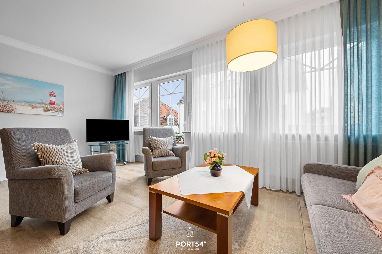 Wohnzimmer mit Sofa, Sesseln, Tisch und TV. Große Fenster mit weißen Vorhängen.