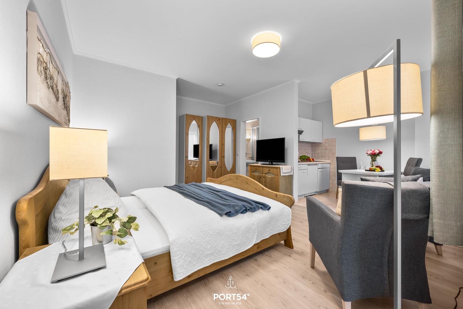 Schlafzimmer mit Bett, Nachttisch und Lampe. Küche und Wohnbereich im Hintergrund.