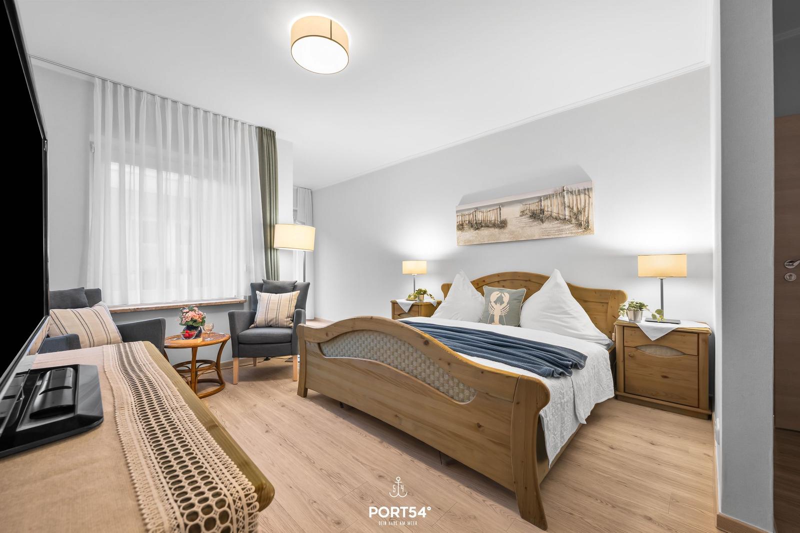 Schlafzimmer mit Holzbett, Nachttischen und Sitzgelegenheit. Fenster mit weißen Vorhängen.