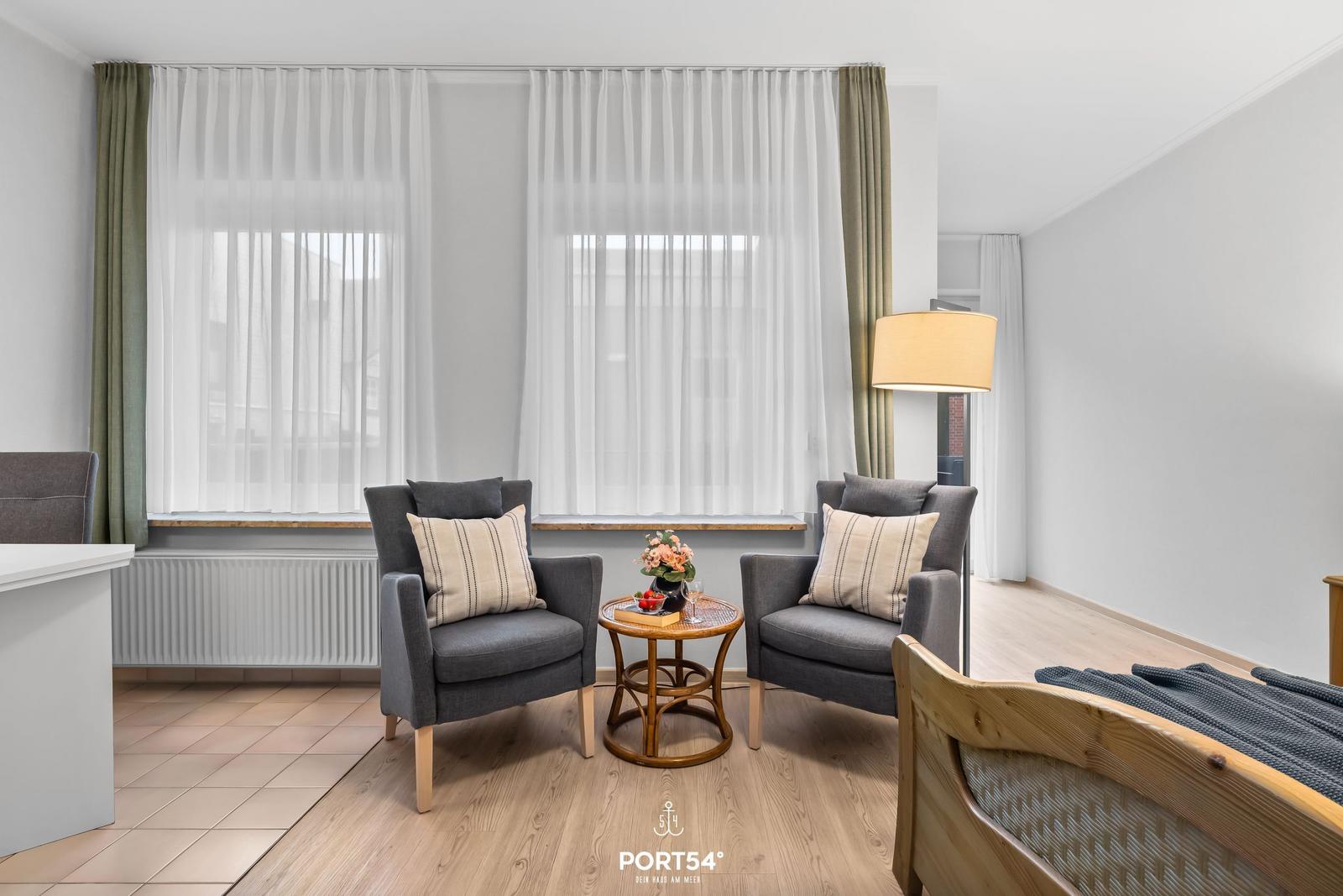 Zwei graue Sessel stehen vor einem Fenster mit weißen Vorhängen. Zwischen ihnen steht ein runder Tisch.