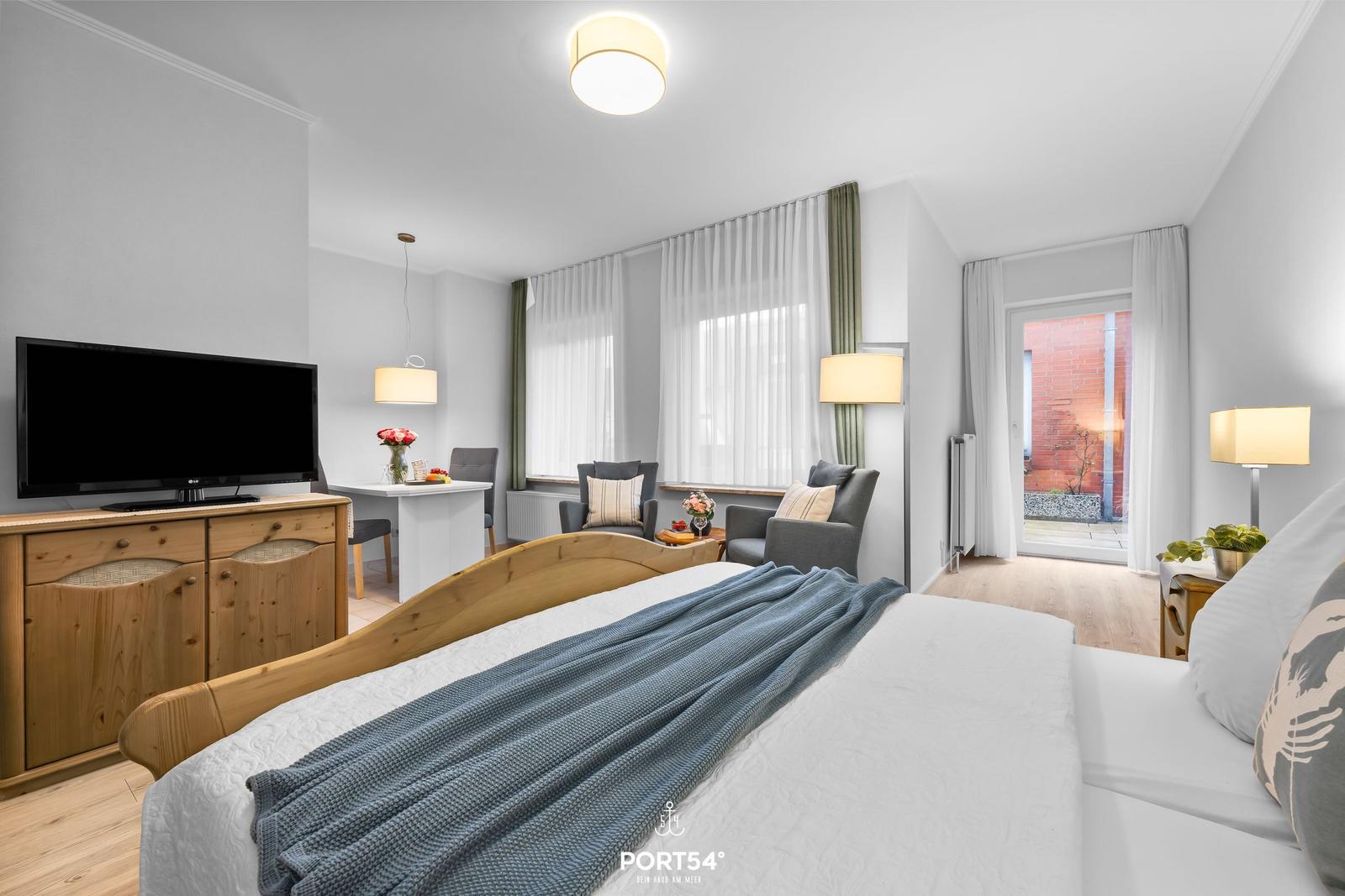 Schlafzimmer mit Bett, TV, Sitzgelegenheit und Balkon. Holzmöbel und Fenster mit Vorhängen.