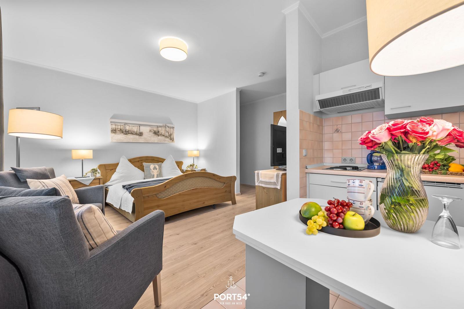 Wohnbereich mit Schlafzimmer und Küche. Auf der Kücheninsel stehen Obst und Blumen.