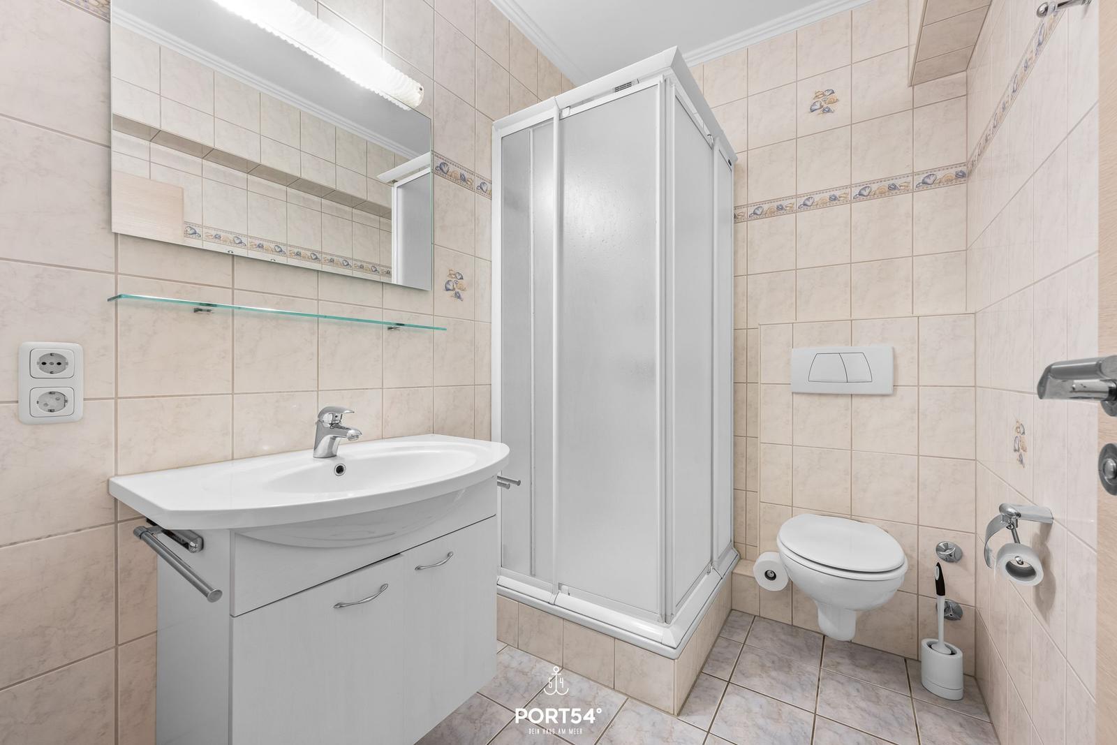 Badezimmer mit Dusche, Waschbecken und Toilette. Fliesenwand mit Muschelrand.