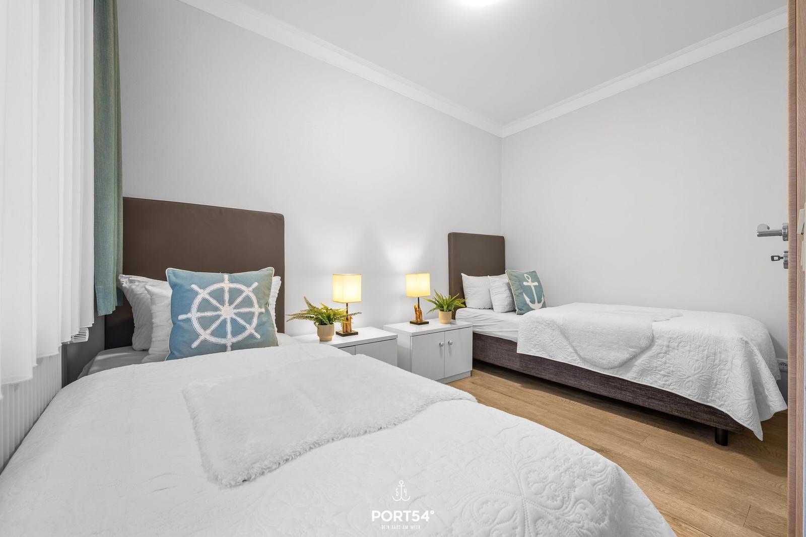 Zwei Betten mit weißen Bettwäschen und maritimen Kissen. Zwischen den Betten steht ein Nachttisch mit Lampen.