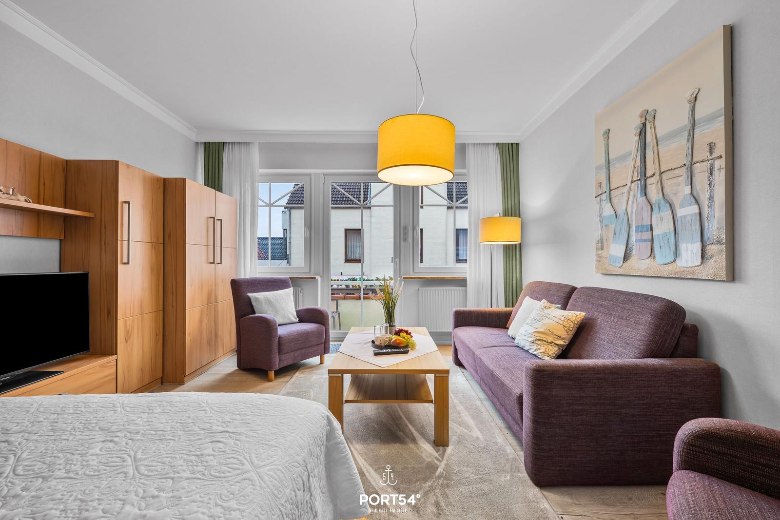 Wohnbereich mit Sofa, Sessel und TV. Große Fenster mit Blick auf Balkon.