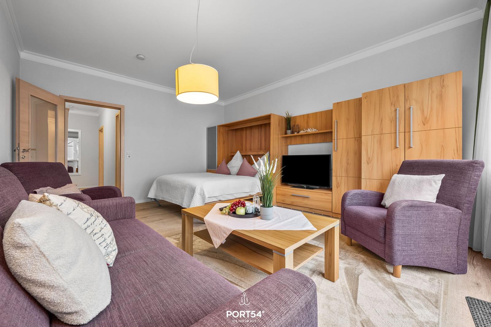 Wohnbereich mit Sofa, Sessel und Bett. Holzmöbel und Fernseher sind sichtbar.