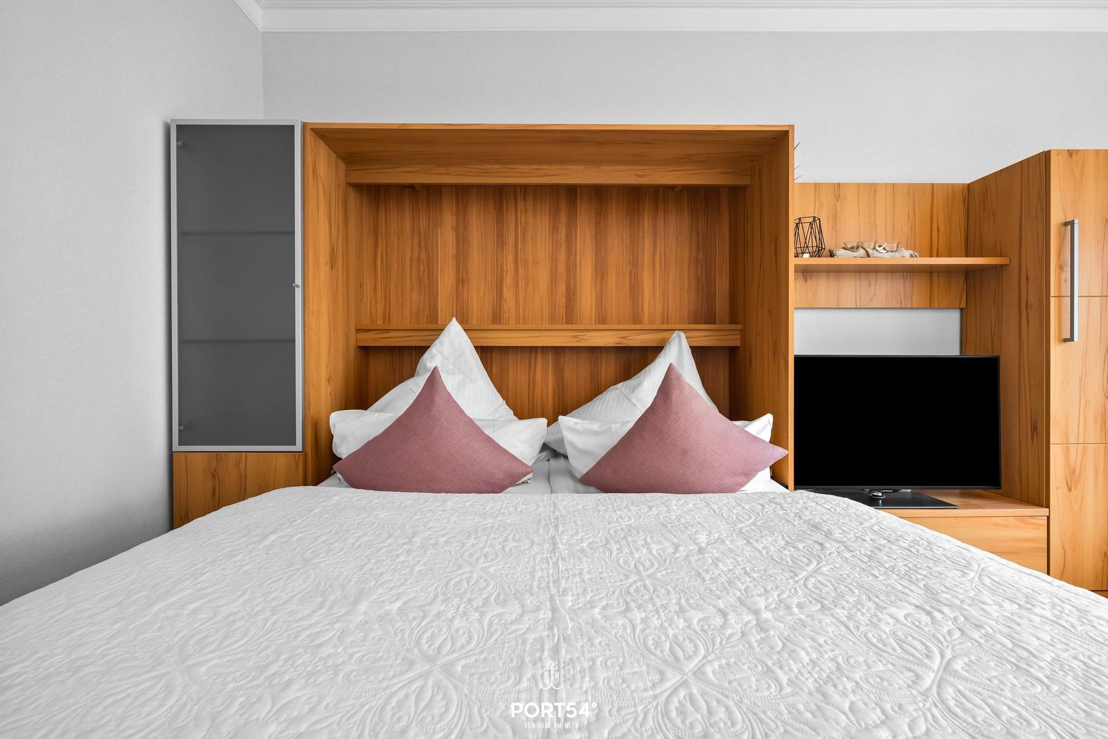 Schlafzimmer mit Bett, Holzregal und Fernseher.