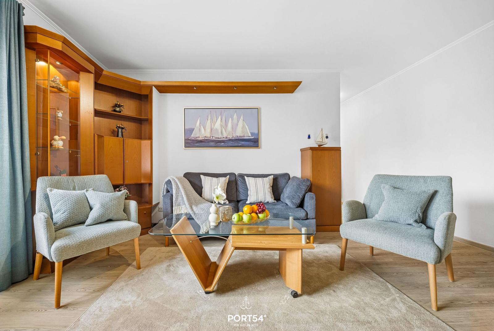 Wohnzimmer mit Sofa, zwei Sesseln und Glas-Tisch. Holzmöbel und Wandbild mit Segelbooten.