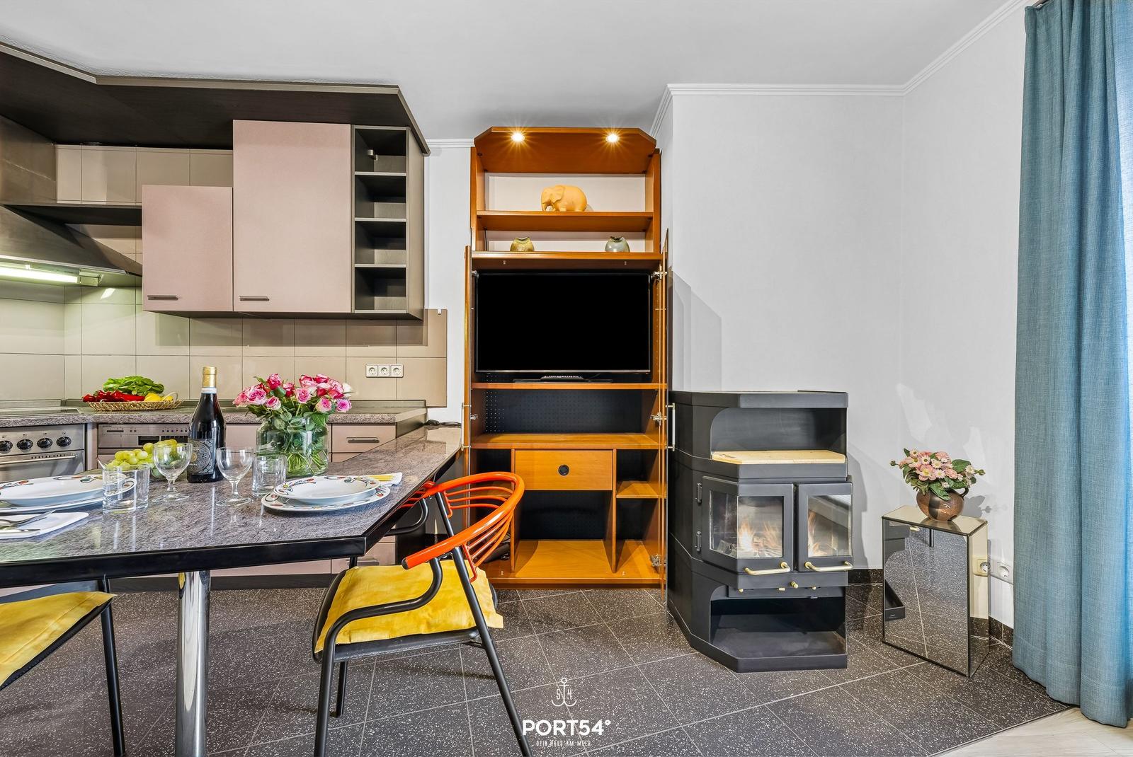 Küche mit Esstisch, TV-Schrank und Kaminofen. Blumen und Wein auf Tisch.