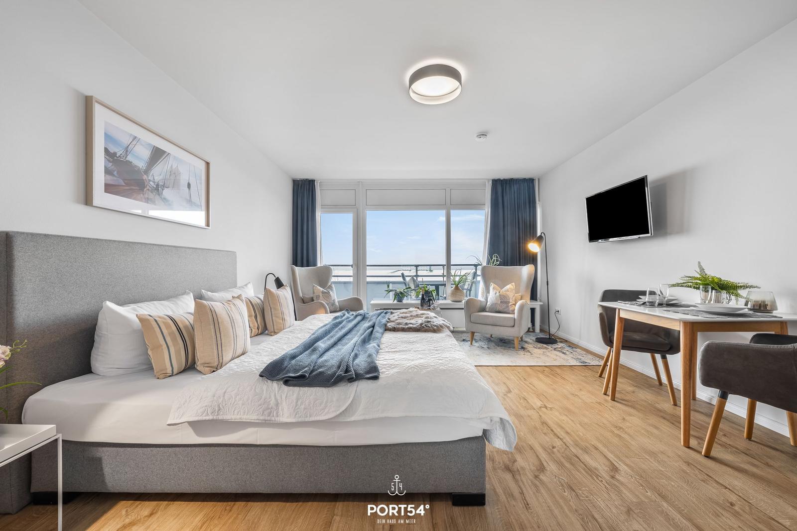 Schlafzimmer mit Bett, Sitzgelegenheiten und Blick auf Balkon. Esstisch mit Stühlen und Fernseher.