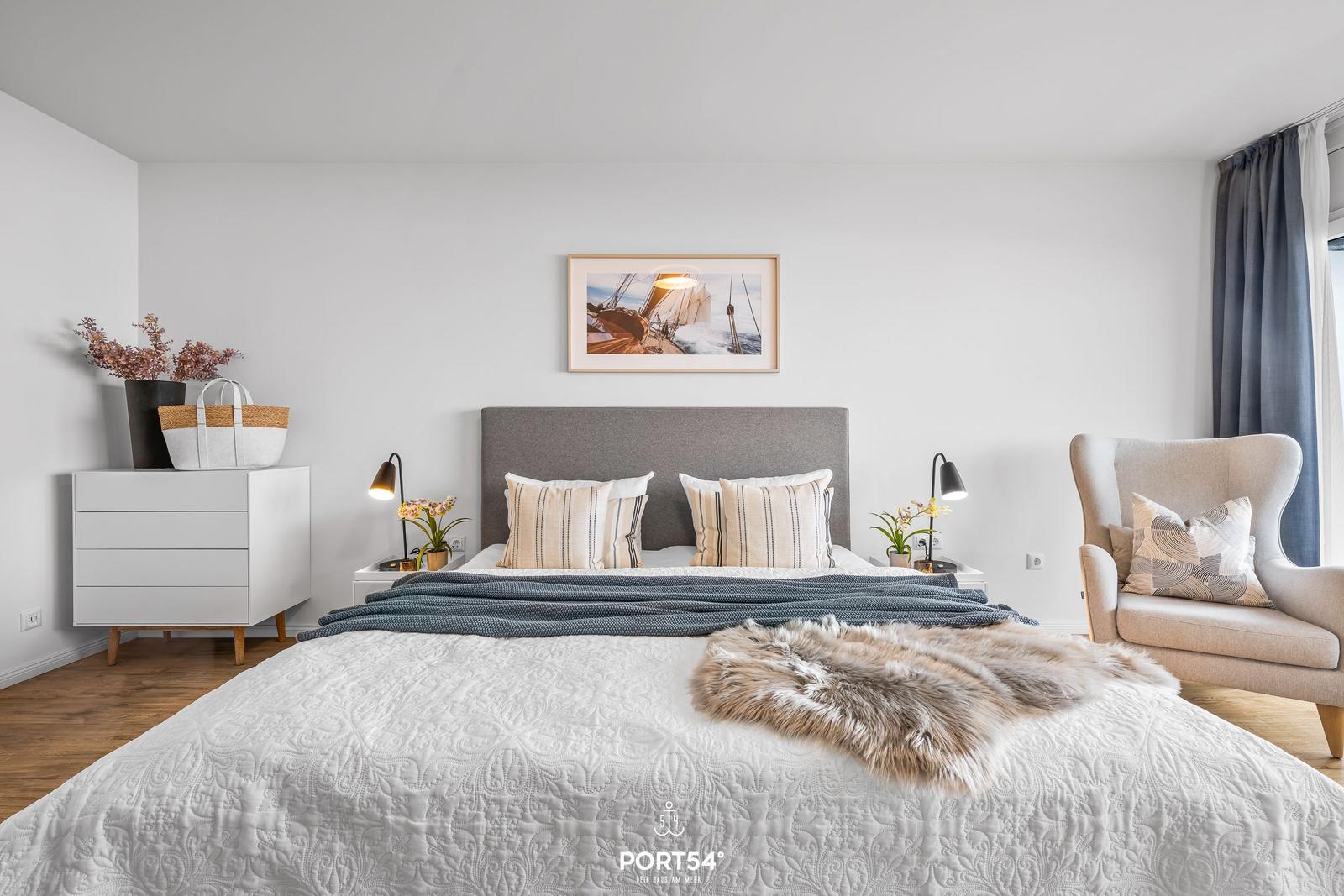 Schlafzimmer mit Bett, Nachttischen, Schrank und Sessel. Wandbild und Vorhänge sind sichtbar.