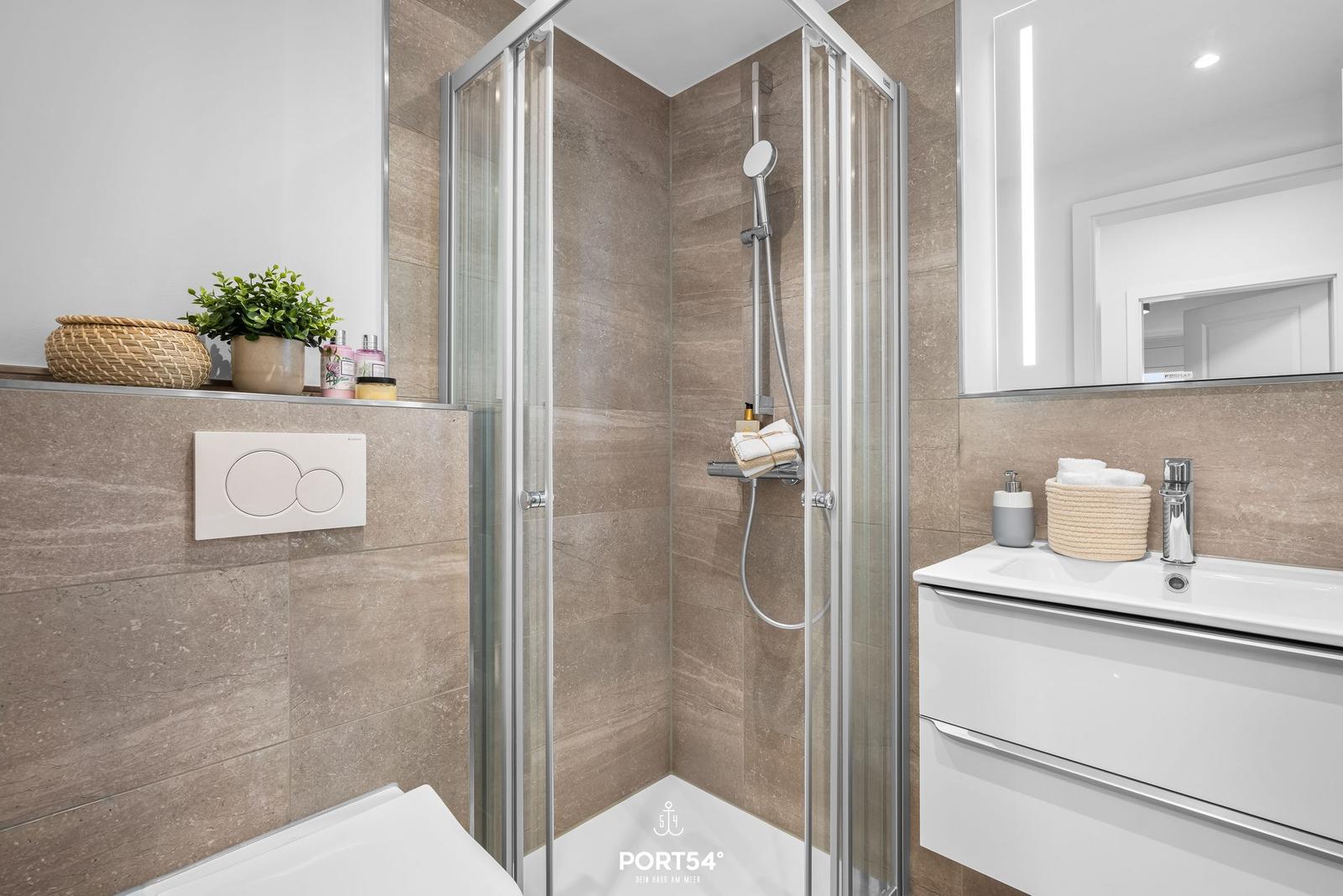 Bad mit Dusche, Waschbecken und Toilette. Fliesenwand und Spiegel mit Beleuchtung.