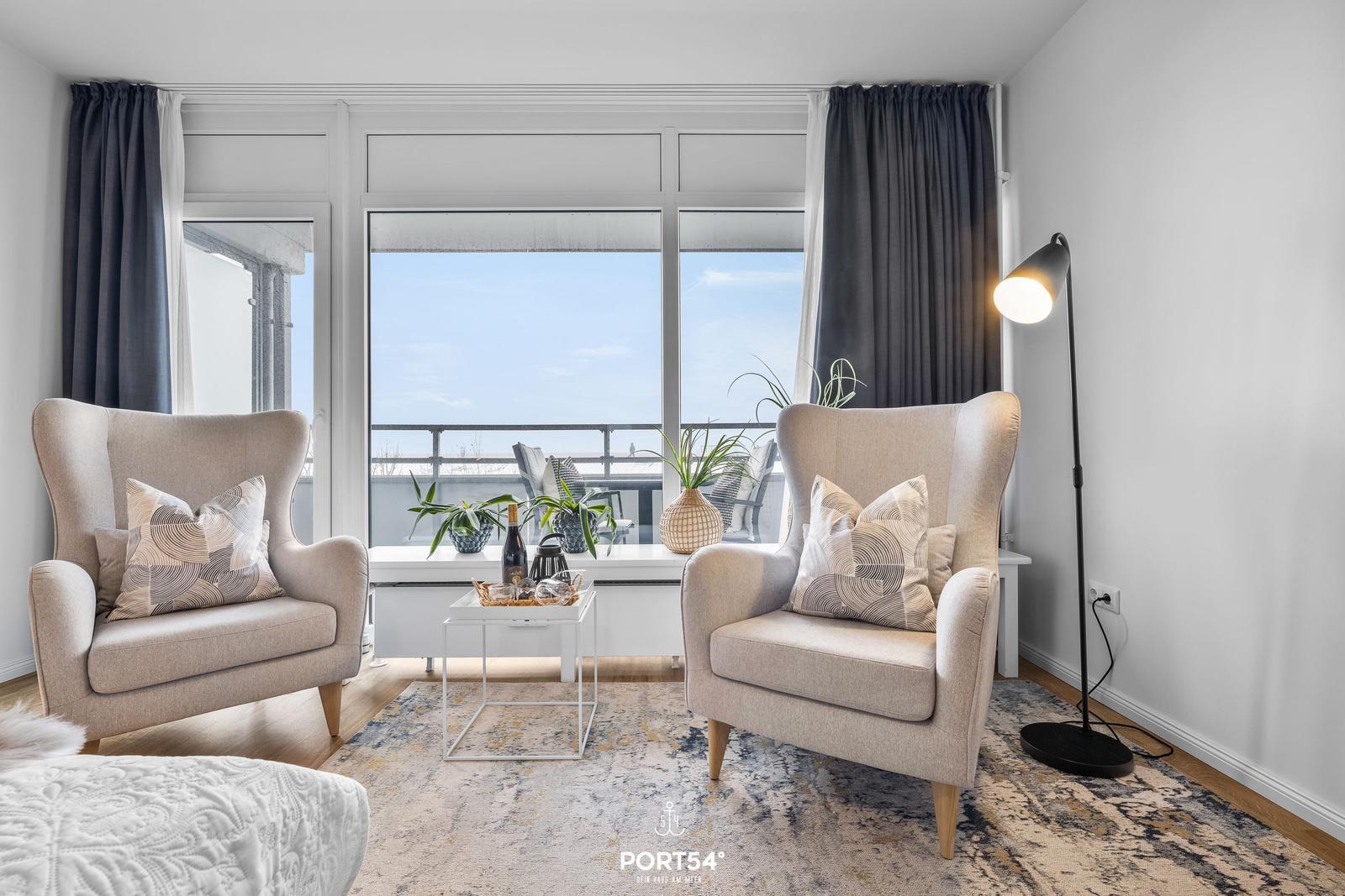 Zwei Sessel stehen vor Fenstern mit Meerblick. Tisch, Teppich und Stehlampe sind vorhanden.