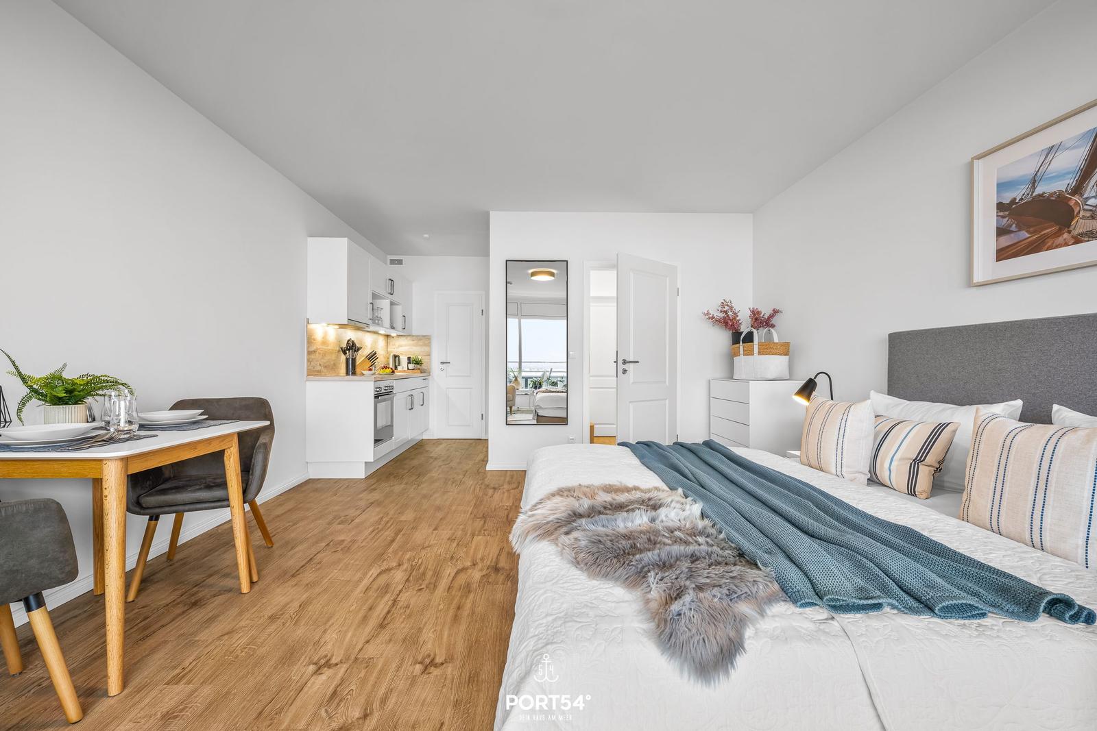 Schlafzimmer mit Bett, Nachttisch und Wandspiegel. Küchenbereich und Esstisch sind sichtbar.