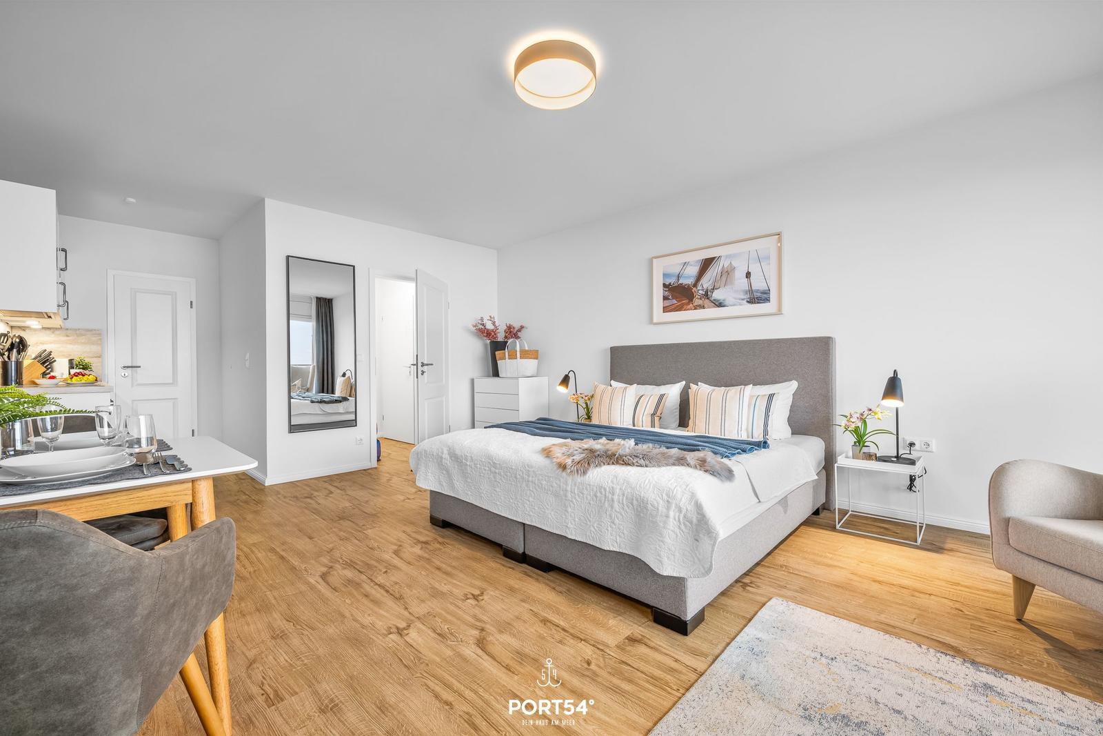 Schlafzimmer mit Bett, Nachttisch und Spiegel. Küchenbereich und Esstisch sind sichtbar.