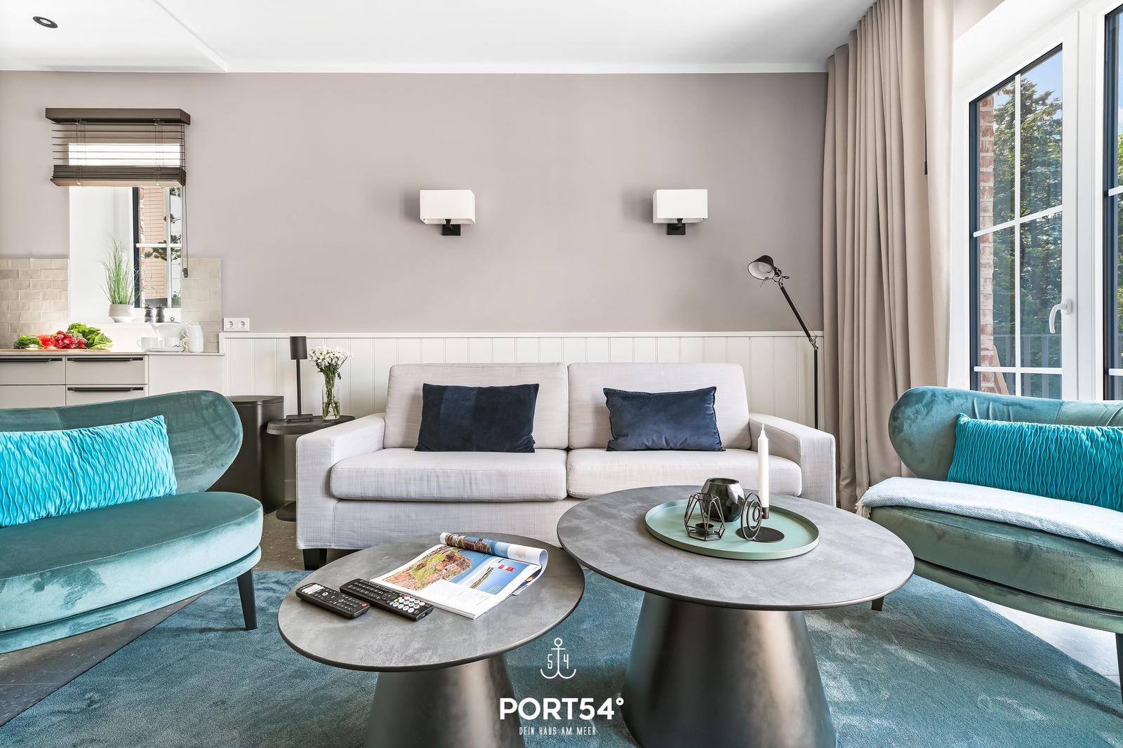 Wohnzimmer mit Sofa, zwei Sesseln und zwei runden Tischen. Durchgang zur Küche und große Fenster.