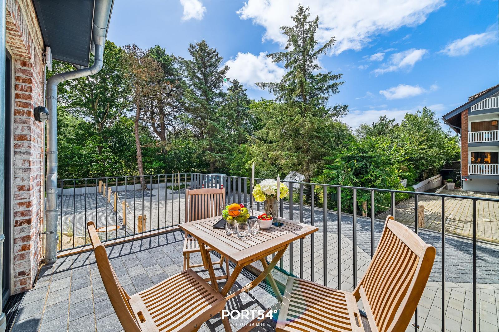 Terrasse mit Holztisch und Stühlen, Blick auf grüne Bäume und einen klaren Himmel.