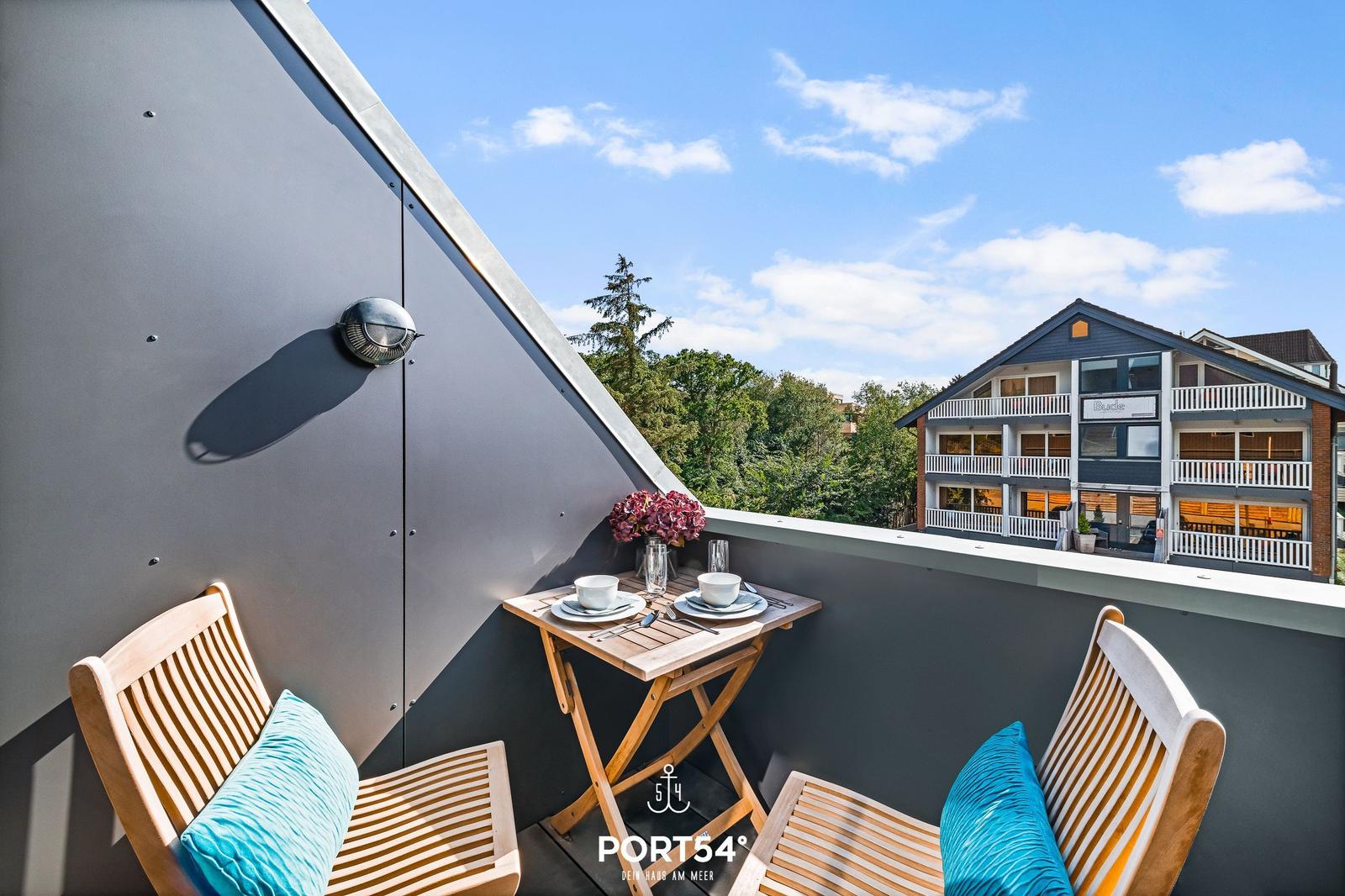 Terrasse mit Tisch und zwei Stühlen, Blick auf ein Gebäude und Bäume.