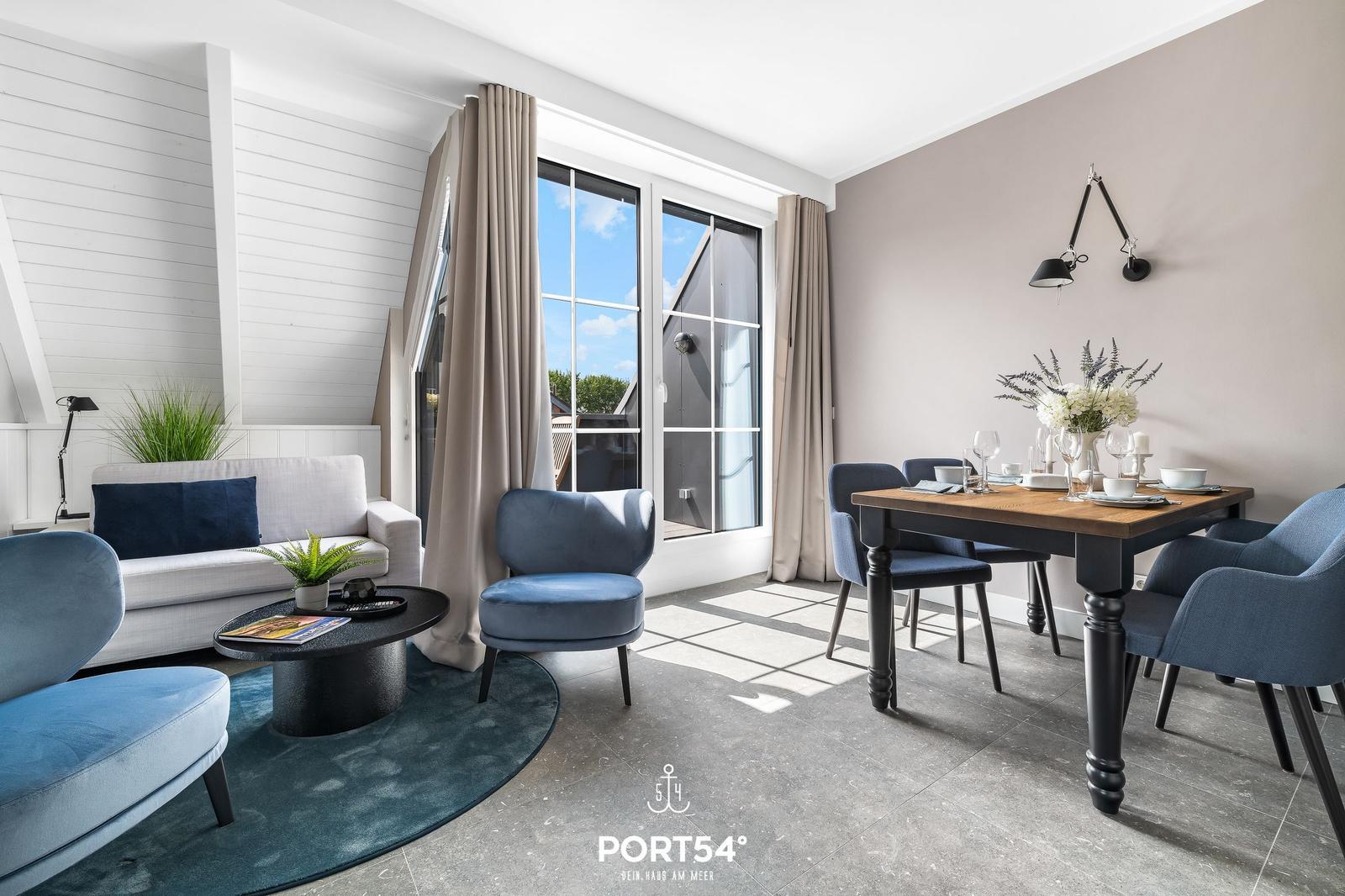 Wohn- und Essbereich mit Sofa, Stühlen und Tisch. Große Fenster mit Blick nach draußen.