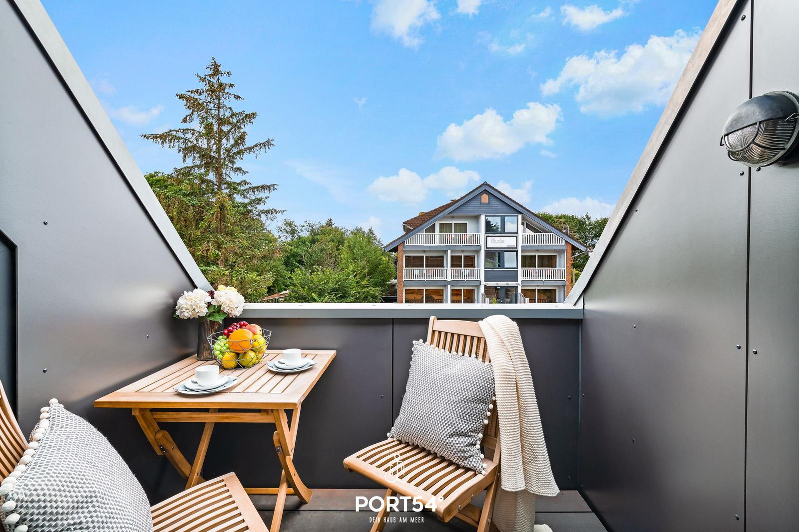 Balcony with wooden table, chairs, and fruit bowl. View of a house and trees under blue sky.