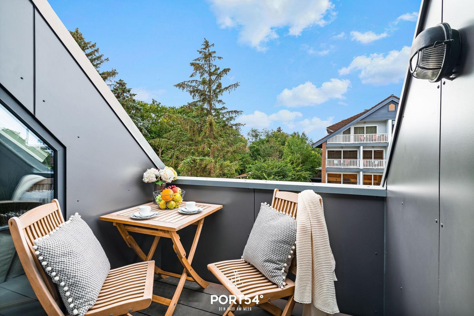 Terrasse mit Tisch, zwei Stühlen, Kissen und Obst. Blick auf grüne Bäume und ein Haus.