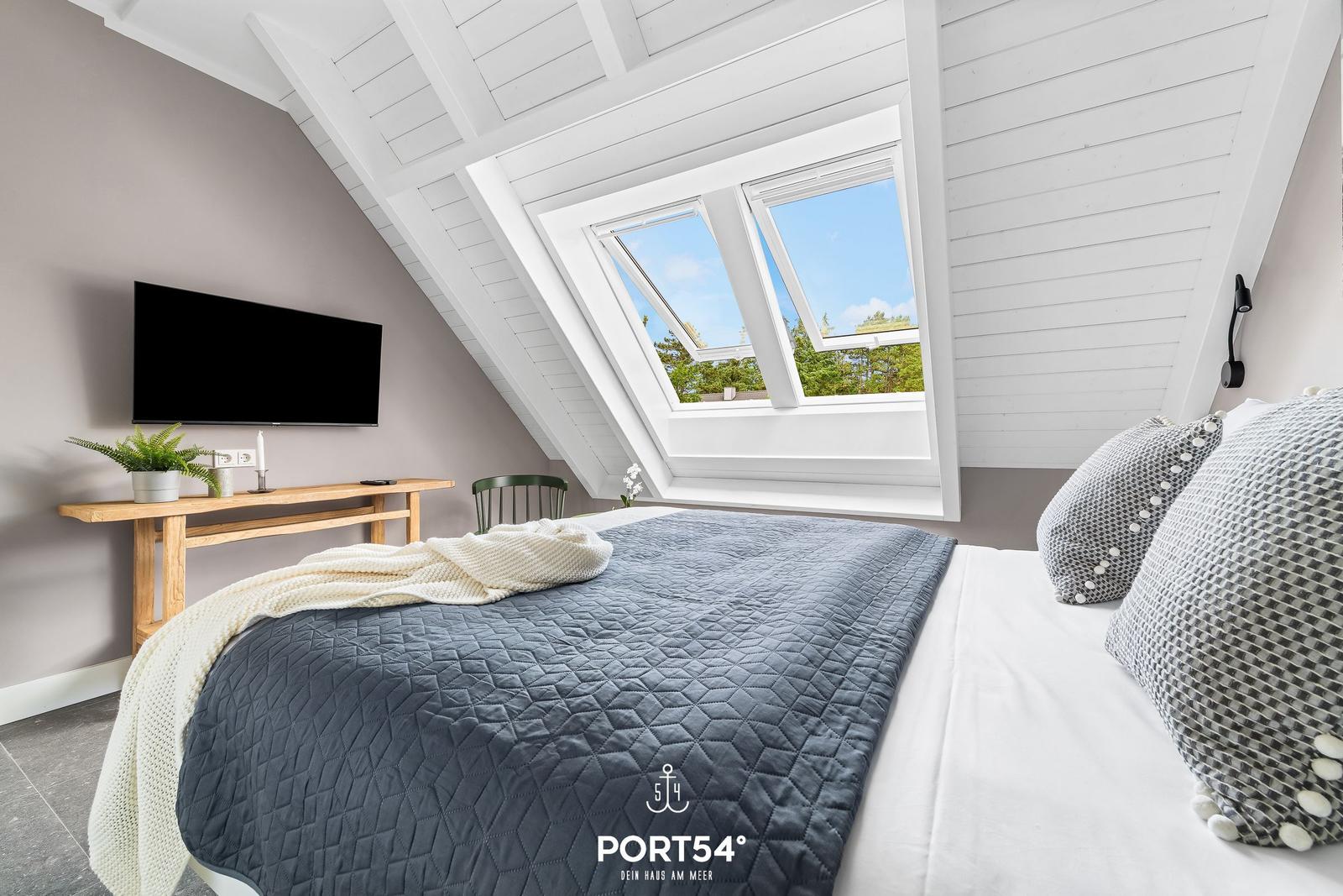 Schlafzimmer mit Dachfenster, Bett, Fernseher und Holztisch.