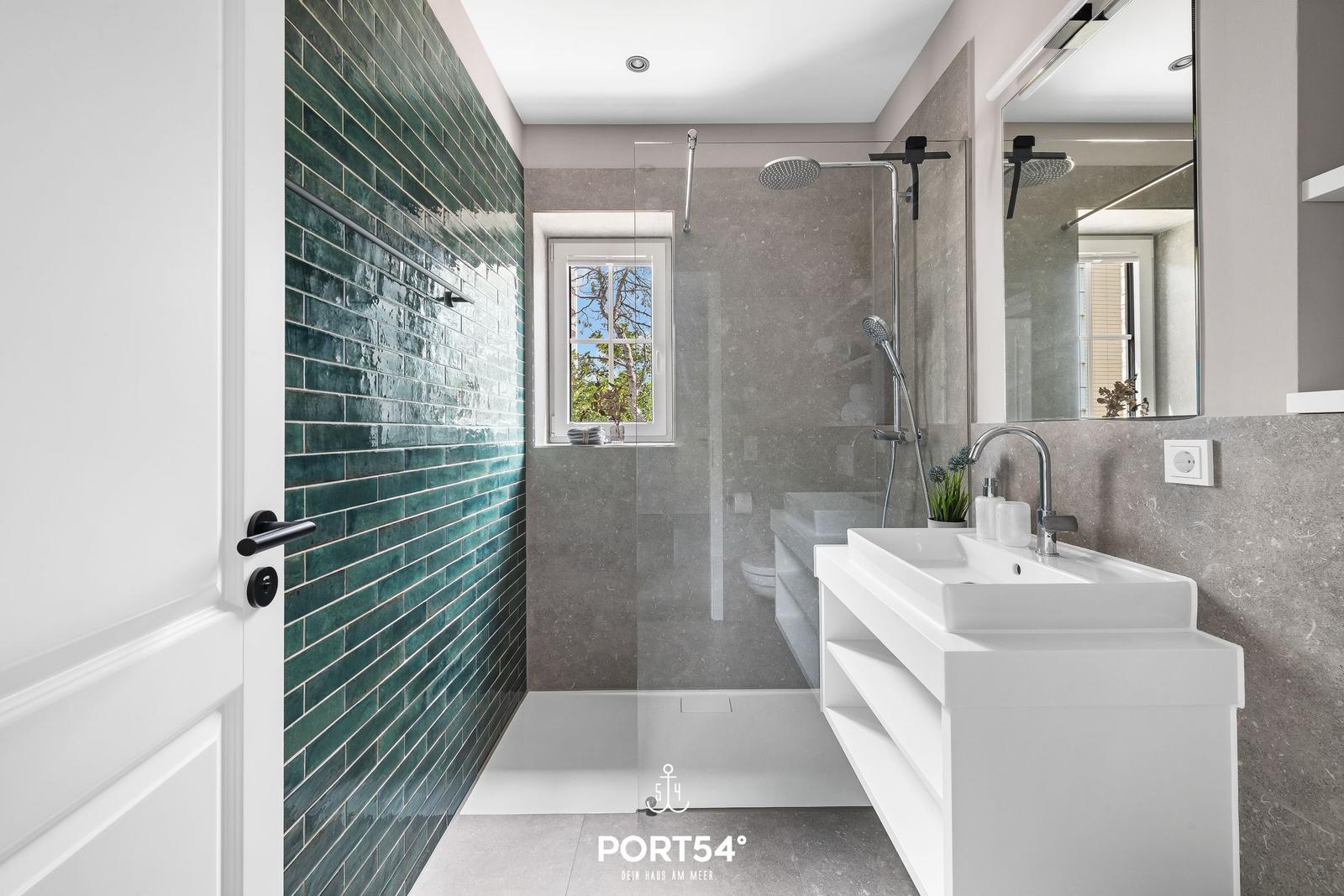 Badezimmer mit Dusche, Waschbecken und grüner Fliesenwand. Fenster mit Blick auf Außenbereich.