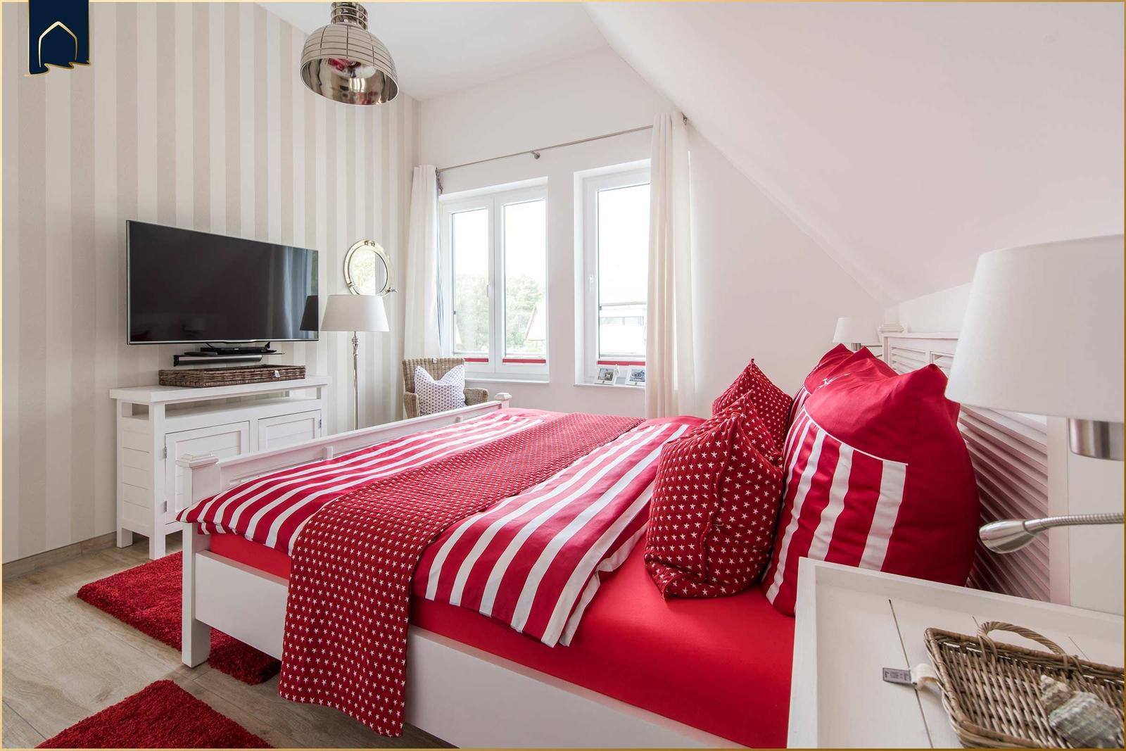 Schlafzimmer mit rotem Bett und TV. Fenster mit weißen Vorhängen. Holzboden und Streifenwand.