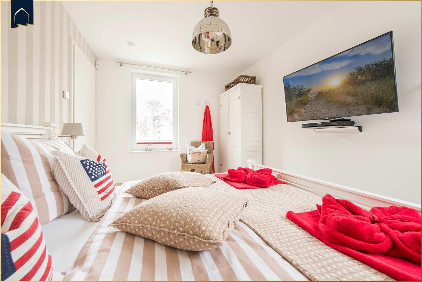 Schlafzimmer mit Bett, TV und Fenster. Rote Decke und Kissen mit US-Flagge.