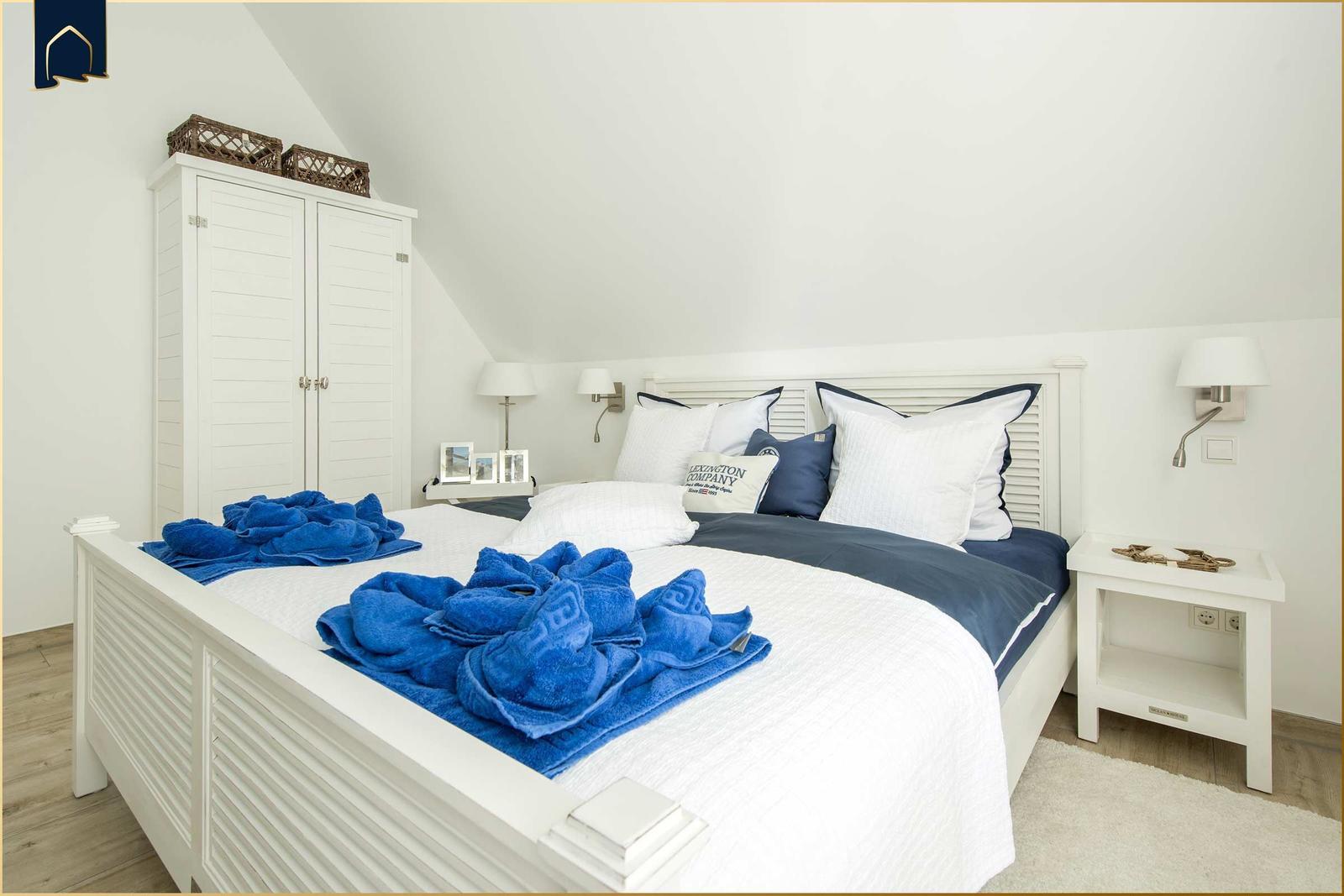 Schlafzimmer mit weißem Bett, blauen Handtüchern und Schrank unter Dachschräge.